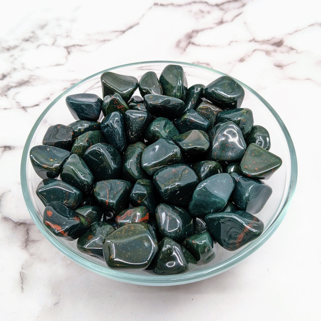 Arafed glass bowl of Bloodstone Green tumbled stones on a marble table