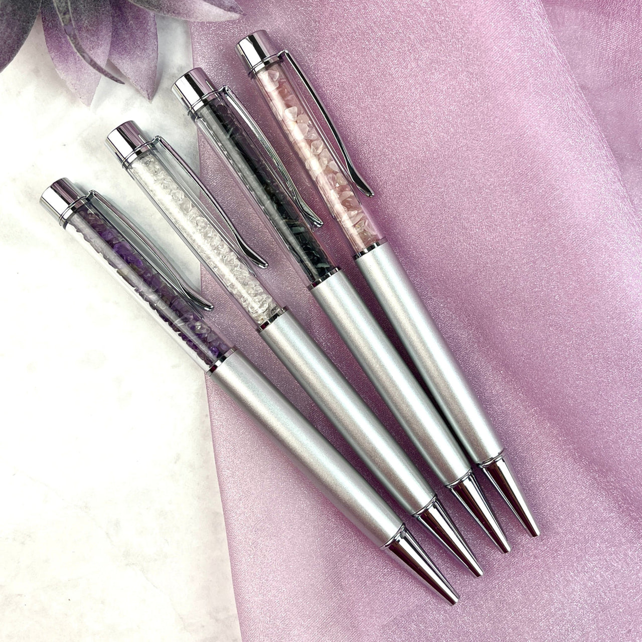 Three rose quartz, clear quartz, and silver tone fountain pens on a purple background