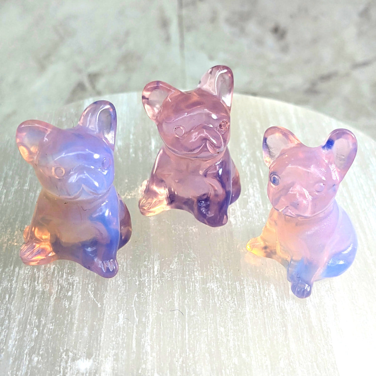 Three pink opalite glass pig figurines on a table from ’Pink Opalite 1.2’ French Bulldog Carving #LV2312’